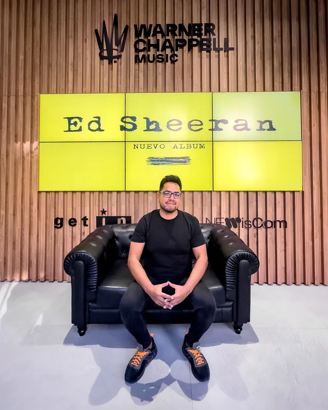 Macuto sentado en un sofá de Warner Chappell Music, bajo el cartel de Ed Sheeran